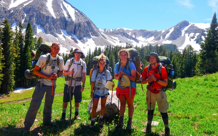 A group of students wearing backpacks pose for a photo in a green meadow. In the background there are snow-capped mountains. 